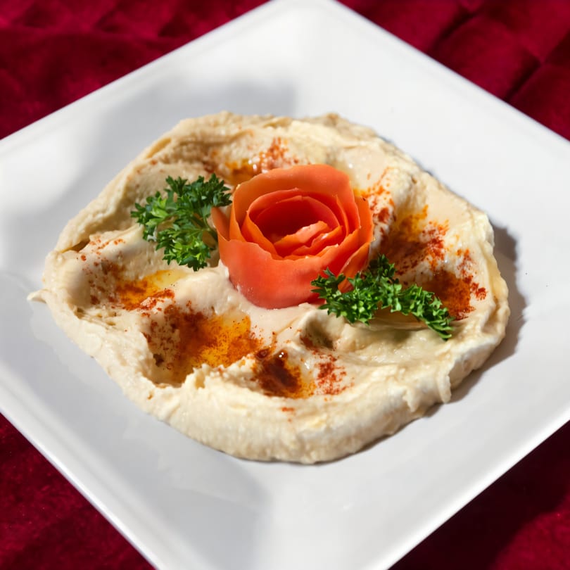 Creamy hummus on a white plate, garnished with a tomato rose, parsley, and paprika.