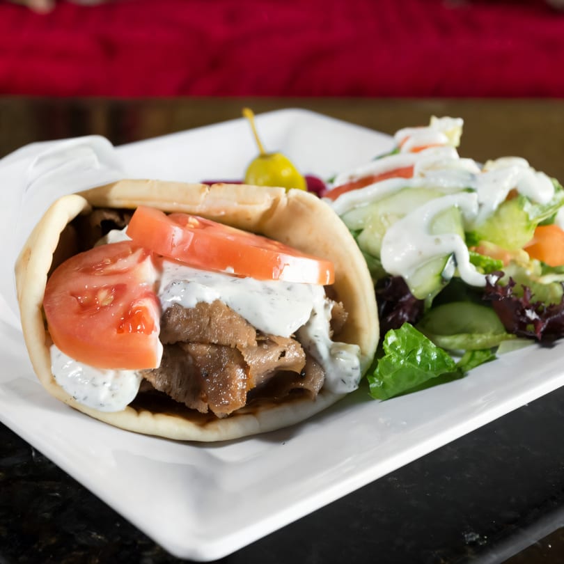 A gyro with meat, tomatoes, and tzatziki sauce, served with a side salad on a white plate.