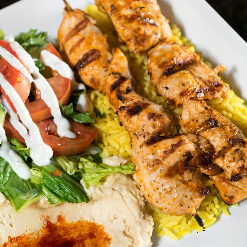 Grilled chicken skewers with yellow rice, hummus, and a fresh salad with dressing.
