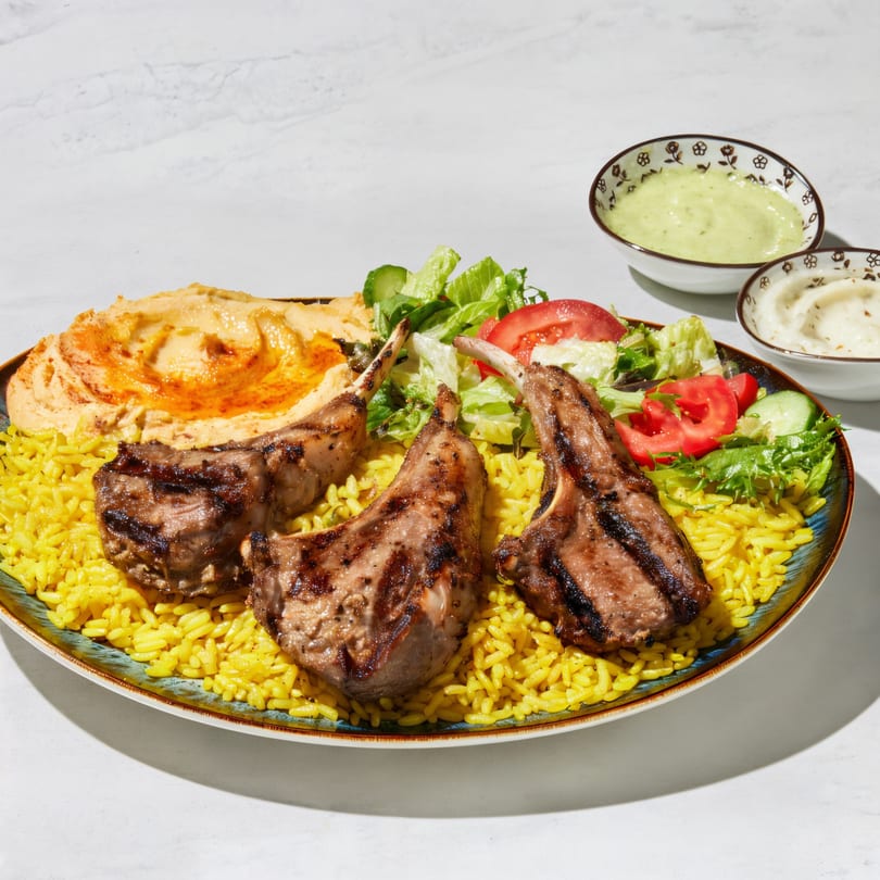 Grilled lamb chops with yellow rice, hummus, salad, and two dipping sauces on a plate.